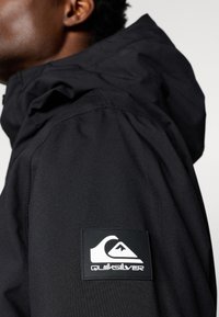 Κοντινό πλάνο ενός ατόμου που φοράει μια μαύρη ζακέτα με λογότυπο Quiksilver στο πάνω μέρος του μπράτσου.