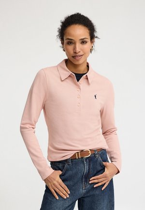 Kvinde iført en lyserød langærmet polo med knapper, en lille broderet logo, blå jeans og et brunt bælte, stående med hænderne i lommerne.