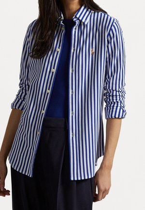 Femme portant une chemise boutonnée à rayures verticales bleues et blanches par-dessus un top bleu foncé et un pantalon noir, avec les manches retroussées jusqu'aux coudes.