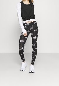 Leggings negros con patrones del logo de Puma en blanco, combinados con una camiseta sin mangas negra y una capa larga de manga blanca. Mujer vistiendo zapatillas blancas.