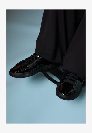 Baskets en cuir verni noir avec des lacets texturés, un design de bout lisse et une semelle noire contrastante. Positionnées sur une surface bleue douce.