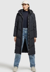 Cappotto lungo imbottito nero con cappuccio, cerniera frontale, marchio bianco sulla cerniera e cuciture evidenti, indossato sopra una maglietta blu e jeans blu.