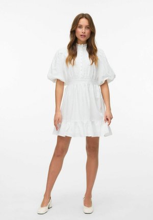 Weißes Kleid mit Puffärmeln, Knopfverschluss vorne und Spitzenakzenten. Ausgestellter Saum mit Rüsche. Getragen mit passenden, hellen Schuhen.