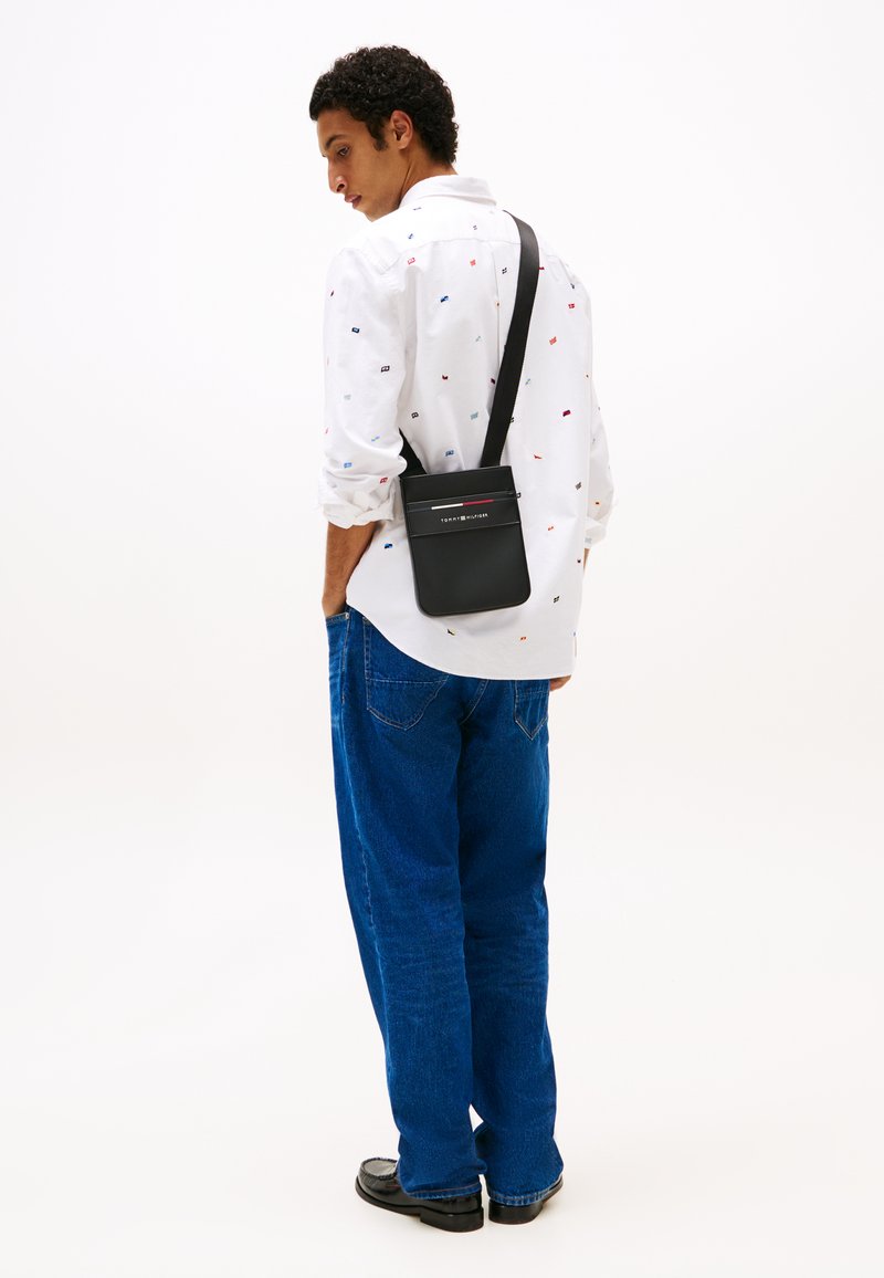 Zwarte leren schoudertas met contrasterende rode en witte strepen, slanke rechthoekige vorm, gedragen over een wit met patroon shirt en blauwe jeans.