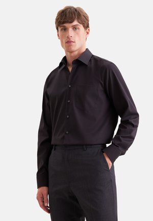 Homme portant une chemise noire à manches longues boutonnée avec une poche poitrine et un pantalon gris foncé, debout avec une main dans la poche.