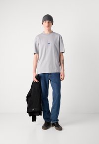 Graues T-Shirt mit kurzen Ärmeln und blauem Logo, blaue Jeans, schwarze Jacke über dem Arm getragen, graue Strickmütze und schwarze Schuhe.