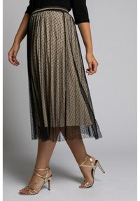 Femme portant une jupe midi beige avec un overlay en maille à pois noirs et des sandales à talons dorées à lanières sur un fond gris.