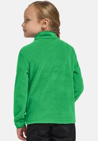Bambina con i capelli intrecciati che indossa una giacca di pile verde brillante e pantaloni neri, rivolta di lato su uno sfondo bianco.