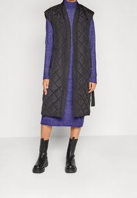 Svart, vadderad ärmlös väst med lång silhuett, bärs över en figurnära lila stickad klänning. Svarta ankelskor fullbordar outfiten.