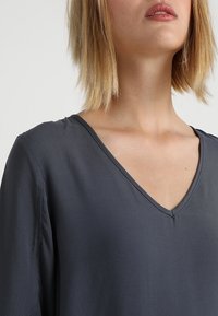 Ciemnoszara bluzka z dekoltem w serek i długimi rękawami, gładka w dotyku, z minimalnymi detalami szycia. Materiał wydaje się lekki i zwiewny.