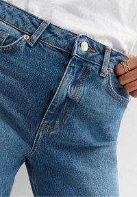 Ljusblå denimjeans med hög midja, framfickor, orange sömmar och en silverknapp vid midjan. Slät textur.