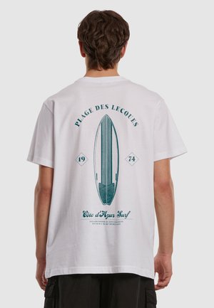 Tânăr purtând un tricou alb cu o grafică teală a unei plăci de surf și textul „Plage des Lecques 1974 Côte d’Azur Surf” pe spate.