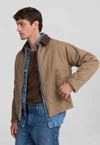 Mężczyzna w warstwowej brązowej kurtce na niebieskiej koszuli dżinsowej i dżinsach, stoi z jedną ręką w kieszeni kurtki, patrząc w lewo.