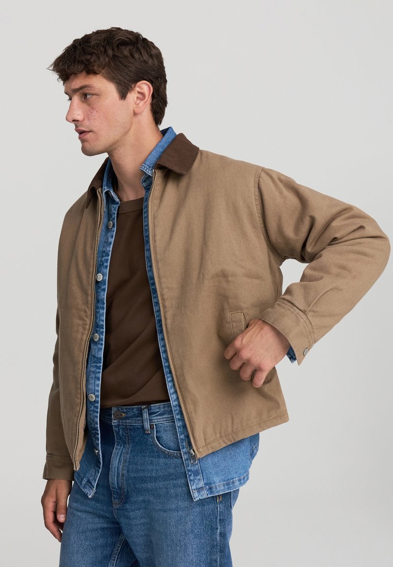 Mężczyzna w warstwowej brązowej kurtce na niebieskiej koszuli dżinsowej i dżinsach, stoi z jedną ręką w kieszeni kurtki, patrząc w lewo.