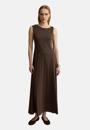 Vrouw met steil blond haar draagt een mouwloze bruine maxijurk en een gouden manchetarmband, gecombineerd met donkerbruine platte schoenen, staand op een witte achtergrond.