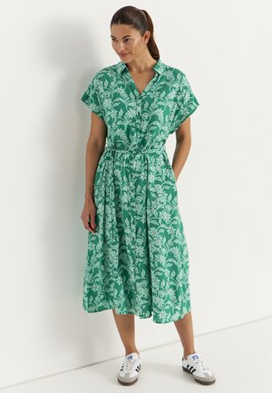 Kvinde står op ad hvid væg iført en grøn, blomsterprintet midi-kjole med knapper, korte ærmer og hvide sneakers.