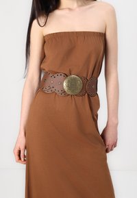 Brązowy skórzany pasek z okrągłą metalową klamrą, z wyciętymi otworami i teksturowanym wzorem, noszony na brązowej sukience bez ramiączek.