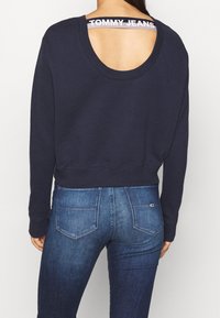 Kvinna iklädd en mörk marinblå sweatshirt med öppen ryggdetalj och "TOMMY JEANS"-rem, kombinerad med blå denimjeans.