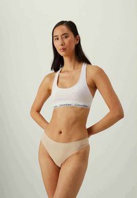 Nais, kes kannab valget Calvin Kleini spordirinnahoidjat ja beeži bikiinistiilis aluspükse, seisab käed selja taga, ühtlasel taustal.