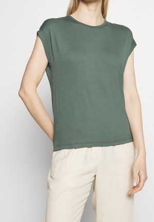 Frau trägt ein olivgrünes Kurzarmtop und hellbeige Hose, steht mit einer Hand in der Tasche und der anderen locker an der Seite.