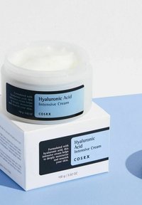 Pot transparent de crème intensive à l’acide hyaluronique COSRX avec étiquette bleu clair sur boîte assortie sur fond bleu et blanc.
