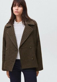 Cappotto doppiopetto a quadri marrone con ampi reverse, tessuto strutturato e dettagli con bottoni, indossato sopra un maglione bianco a maglia. Jeans scuri visibili.