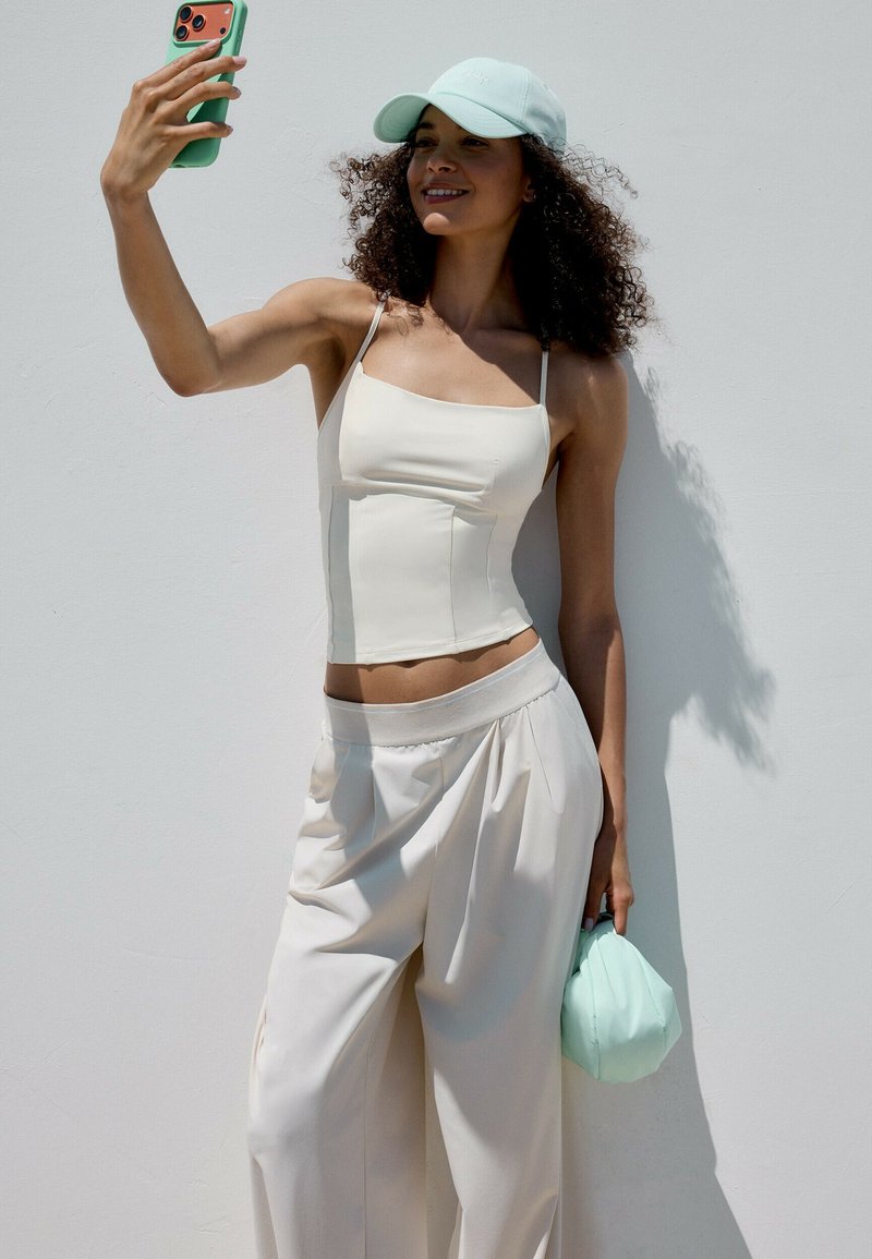 Femme portant un haut blanc, un pantalon large et une casquette bleu clair prend un selfie avec son téléphone, tenant un sac souple bleu clair assorti à ses côtés.