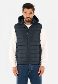 Gilet imbottito navy con cappuccio, caratterizzato da un motivo a coste orizzontali, chiusura frontale con zip e tasche laterali, indossato sopra una camicia bianca.
