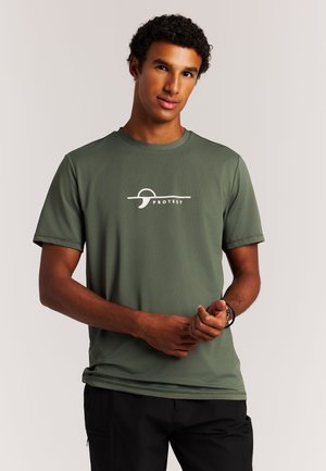 Man draagt een olijfgroen T-shirt met korte mouwen en een wit "Protest"-logo, staat tegen een effen lichte achtergrond, handen voor zich gevouwen.