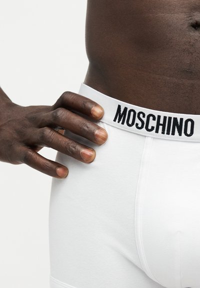 Mørkhudet person iført hvide Moschino boxershorts, hånd hvilende på hoften, synlig mave og linning med logo på ensfarvet baggrund.