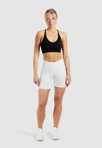 Schwarzer Sport-BH mit gerafften Details und weiße, hoch taillierte Radlerhose. Model trägt weiße Sportschuhe und steht vor einem neutralen Hintergrund.