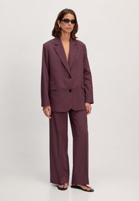 Paarse pak met een lange blazer met gekartelde revers, twee knopen en zijzakken, gecombineerd met wijde broekspijpen. Minimalistisch ontwerp.