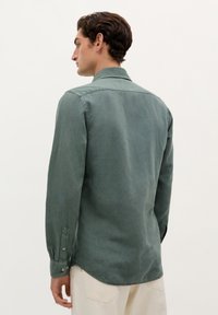 Camisa de manga larga en tela verde apagado, con un collar clásico, puños abotonados y un dobladillo curvado, vista desde la parte de atrás.