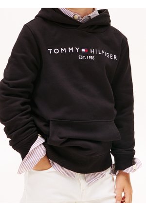 Czarna bluza z kapturem z białym wyszywanym logo "TOMMY HILFIGER EST. 1985", posiadająca przednią kieszeń i ściągacze, noszona na paskowaną koszulę.