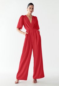 Kvinde iført en rød wide-leg jumpsuit med en dyb V-udskæring og flagrende ærmer, stående med hænderne i lommerne på en hvid baggrund.