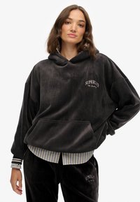 Sudadera con capucha de terciopelo negro con un bolsillo frontal, con "SUPERDRY" bordado en blanco. Combinada con una camisa a rayas azul debajo.