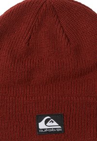 Bonnet en tricot rouge avec une texture côtelée et un couronne plate. Présente un patch logo carré noir avec un graphique de vague blanche sur le devant.