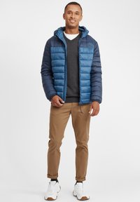 Blå pufferjakke med horisontalt quiltet design, hette og logo-lapp; lagd over en mørk genser; beige bukser og hvite sneakers.