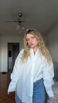 Fehér gombos ing hosszú ujjakkal, túlméretezett fazon; világoskék farmernadrággal párosítva; hullámos haj és világos háttér látható.