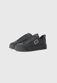 Black sneakers with textured leather upper, flat rubber sole, round toe, and lace-up front. Features a small rectangular branding accent.