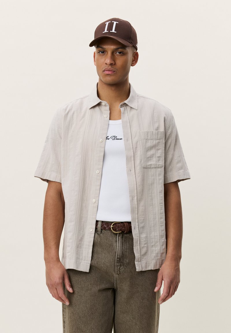 Homme portant une casquette marron, une chemise beige à manches courtes boutonnée sur un débardeur blanc, une ceinture marron et un pantalon vert olive sur fond uni.