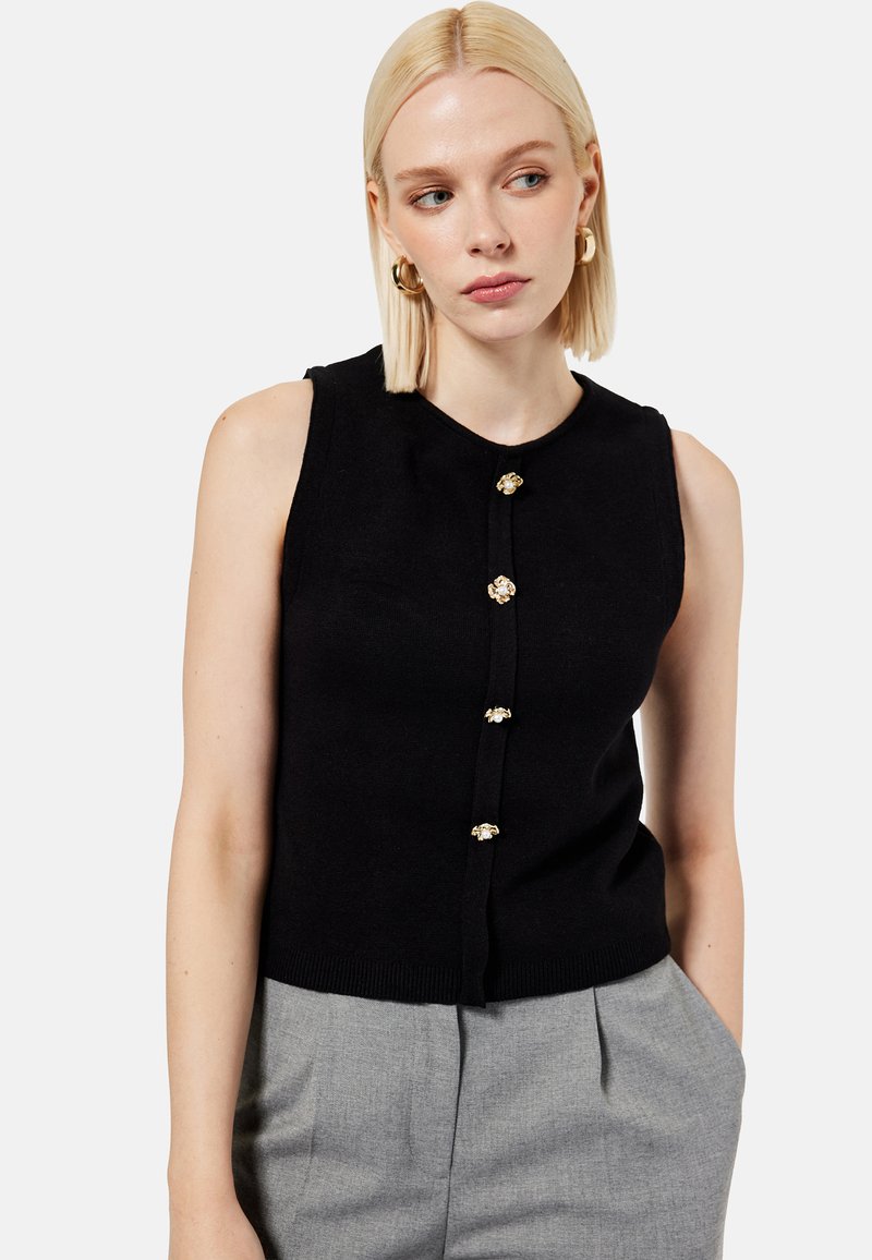 Femme blonde portant un haut noir sans manches avec des boutons en forme de fleurs dorées et un pantalon gris, regardant vers sa gauche.