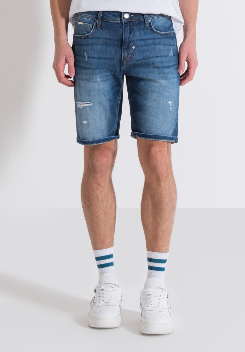 Shorts in denim di un azzurro chiaro con effetti di usura e orli arrotolati, abbinati a scarpe da ginnastica bianche e calze a righe blu e bianche.
