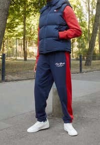 Granatowa kamizelka puchowa noszona na czerwonej koszulce z długim rękawem, zestawiona z granatowymi spodenkami dresowymi z czerwonymi bocznymi panelami i nadrukiem „New York”, oraz białymi trampkami.