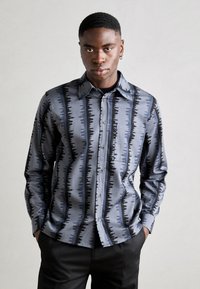 Camisa de manga longa azul e preta com um padrão de ondas, feita de tecido suave. Apresenta um colarinho e fecho com botões, usada com calças escuras.