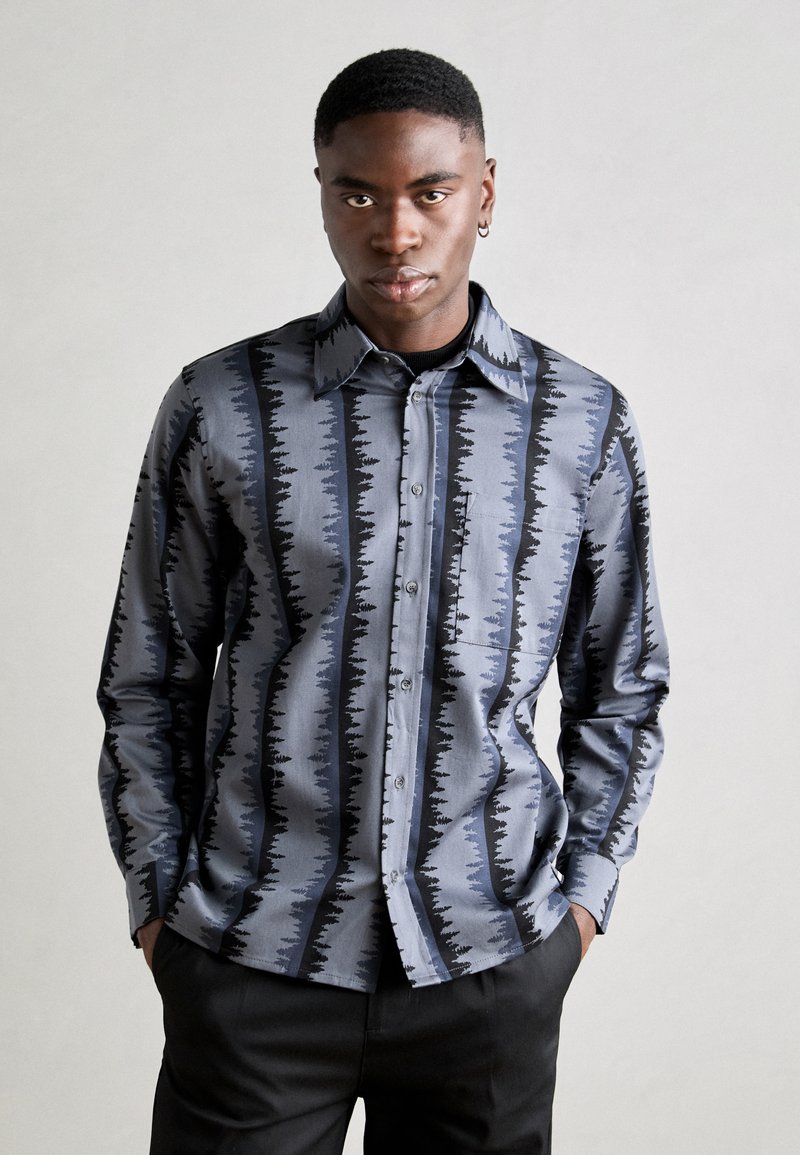Camisa de manga longa azul e preta com um padrão de ondas, feita de tecido suave. Apresenta um colarinho e fecho com botões, usada com calças escuras.