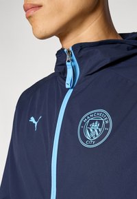 Marineblauwe zip-up jack met lichtblauwe accenten; heeft het Manchester City-logo en het Puma-logo op de borst en schouder; gladde textuur.