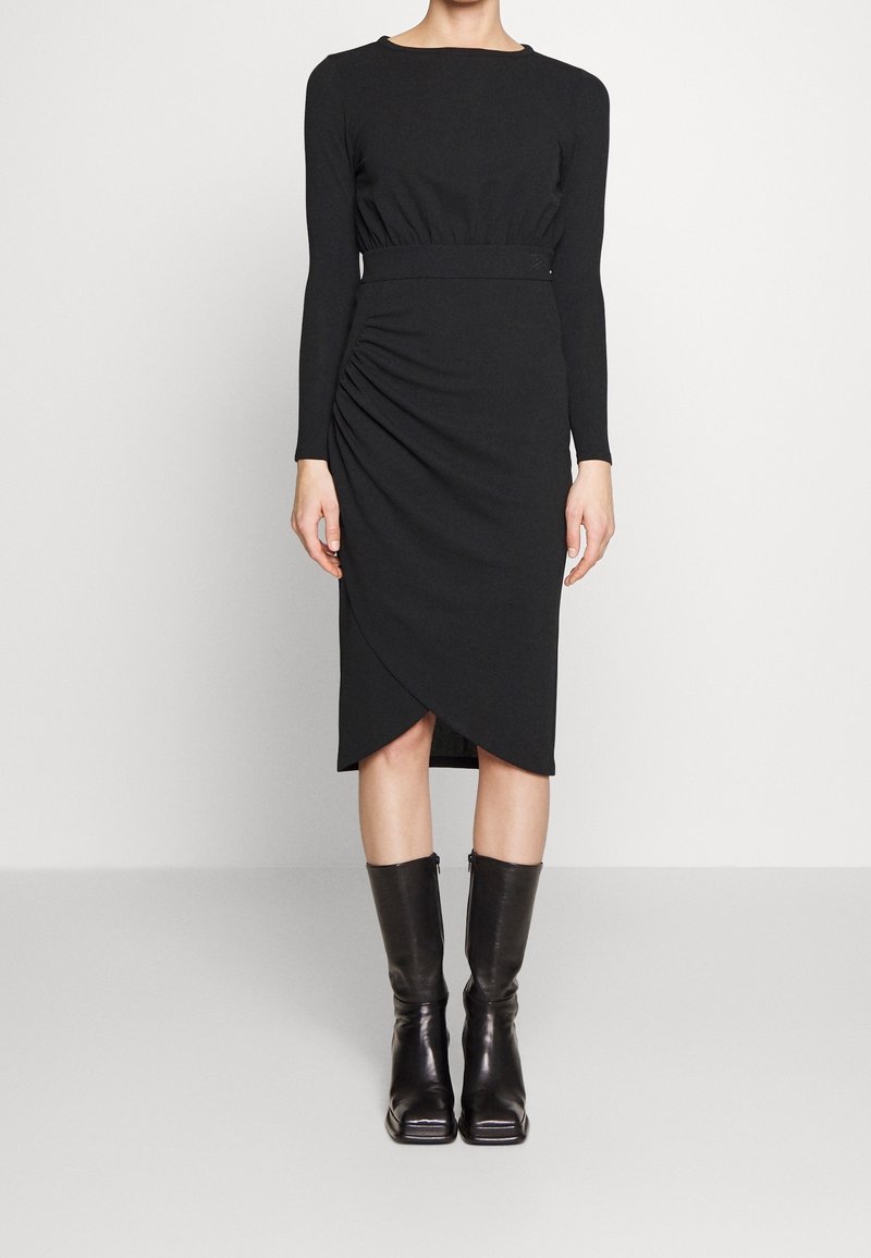 Robe noire à manches longues avec des détails froncés sur le côté et un design de fausse encolure. Fabriquée dans un tissu lisse, associée à des bottes noires jusqu'aux genoux.