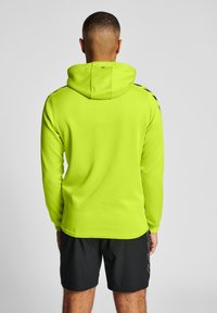 Kapuzensweatshirt in leuchtendem Neon-Gelb, mit glatter Textur und schwarzen Akzenten auf den Schultern. Kombiniert mit schwarzen Shorts.