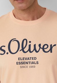 Lichte perzikkleurige T-shirt van katoen, met een groot donkerblauw gedrukt logo en tekst in een casual lettertype. Met een ronde halsontwerp.